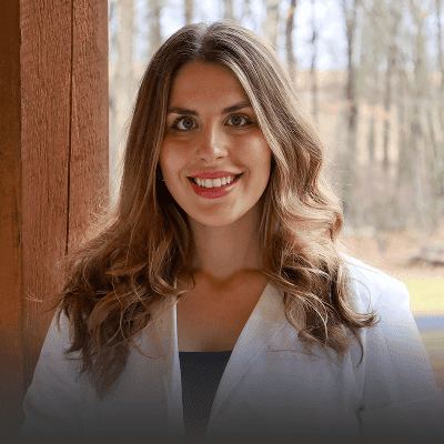A woman with long wavy brown hair, wearing a white coat, stands outside near a wooden post with trees and a blurred background behind her, representing the caring spirit of Grow With Me Pediatrics.