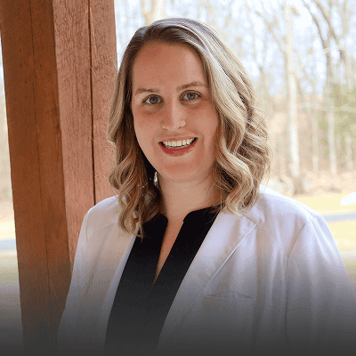 A woman with shoulder-length wavy blonde hair, wearing a white coat over a black top, stands outdoors by a wooden post with trees in the background, representing Grow With Me Pediatrics.