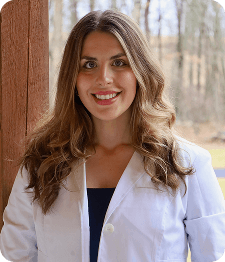 A woman with long brown hair wearing a white lab coat stands outside in front of a wooden post, with trees and grass visible in the background, representing Grow With Me Pediatrics.