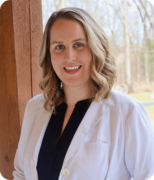 A woman with shoulder-length wavy hair wearing a white coat and dark top stands outdoors near a wooden post, smiling at the camera, representing Grow With Me Pediatrics.