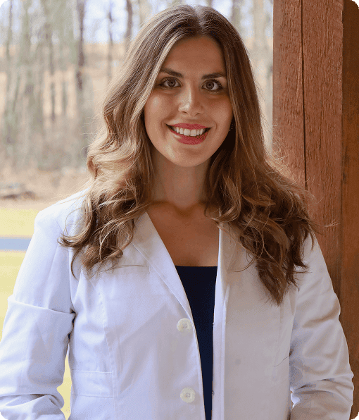 A woman with long brown hair, wearing a white lab coat and dark top, stands outside in front of a wooden post with trees and grass in the background, representing Grow With Me Pediatrics.