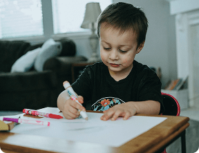 A young child sits at a table, drawing on white paper with markers—a joyful moment supported by the nurturing care at Grow With Me Pediatrics.