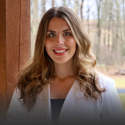 A woman with long wavy hair wearing a white coat stands outdoors in front of a wooden post and trees, smiling at the camera.
