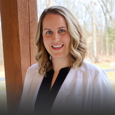 A woman with shoulder-length wavy blond hair, wearing a white coat over a black top, stands outside in front of a wooden post with trees in the background.