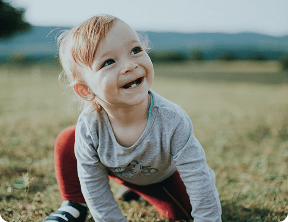 A young child with light hair and missing front teeth crouches on grass outdoors, smiling and looking up, with a blurred landscape in the background—capturing the joyful spirit nurtured at Grow With Me Pediatrics.