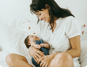 Woman sitting on a bed, smiling and holding a baby in her arms, both facing each other in a bright room—capturing a heartwarming moment inspired by Grow With Me Pediatrics.
