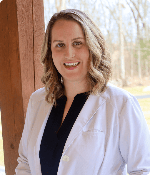 A woman with shoulder-length blonde hair, wearing a white lab coat over a dark top, stands outdoors and smiles at the camera, representing Grow With Me Pediatrics.
