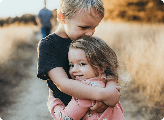 A young boy hugs and kisses the forehead of a smiling young girl on a path through a dry, grassy field. An adult is visible in the blurred background, capturing a heartwarming moment perfect for Grow With Me Pediatrics.