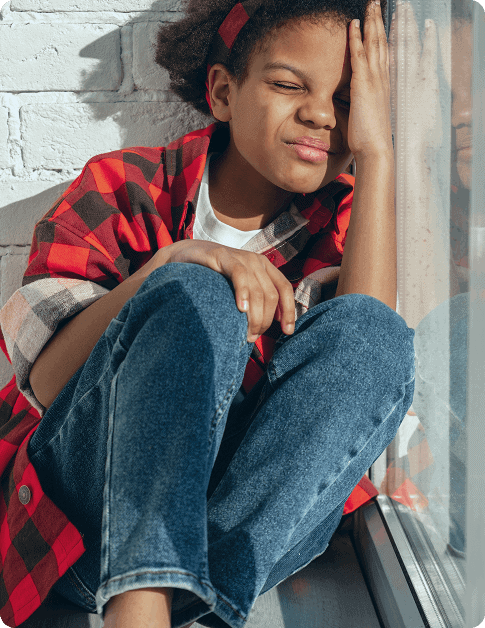 A child in a red plaid shirt and blue jeans sits by a window, squinting and shielding their eyes from the sunlight—a bright moment captured at Grow With Me Pediatrics.