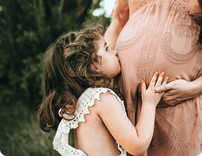 A young girl kisses and touches the belly of a pregnant woman in a lace dress, standing outdoors with greenery in the background, capturing a tender moment celebrated by Grow With Me Pediatrics.