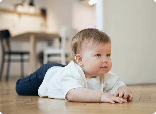 A baby lies on their stomach on a wooden floor, looking forward with arms extended. A blurred table and chairs are visible in the background, capturing a sweet moment of early development at Grow With Me Pediatrics.