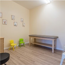 A small medical examination room with an exam table, two child-sized chairs, a stool, and framed pictures on the wall.