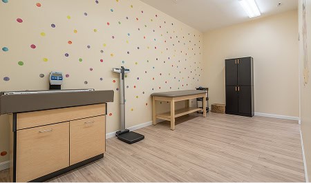 A pediatric exam room with a changing table, digital scale, exam table, cabinet, and colorful polka dots on the wall.