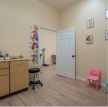 A pediatric exam room with light wood flooring, beige walls, a pink child’s chair, medical equipment, and children’s wall art. An open door reveals another decorated room with a scale.